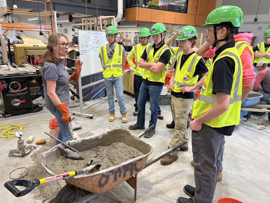 A woman with a tool is pointing at cement in a wheelbarrow while others watch.
