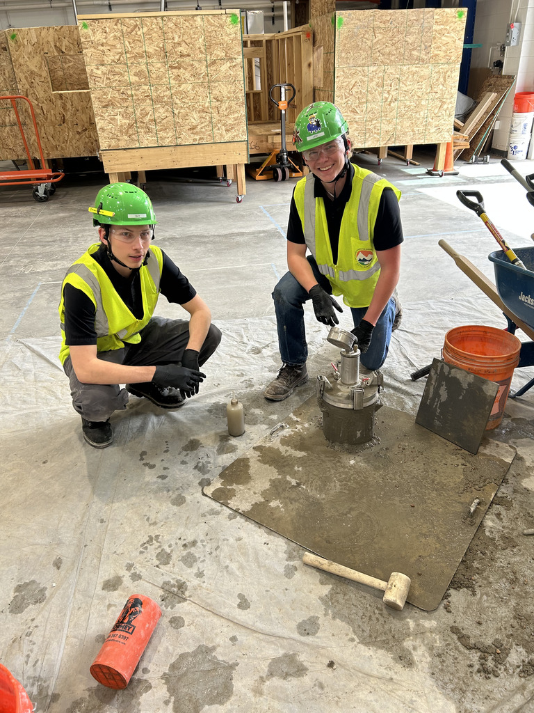 Two workers in protective gear, kneeling, use tools on a cement platform. Tools, a wheelbarrow, and a cart are nearby.