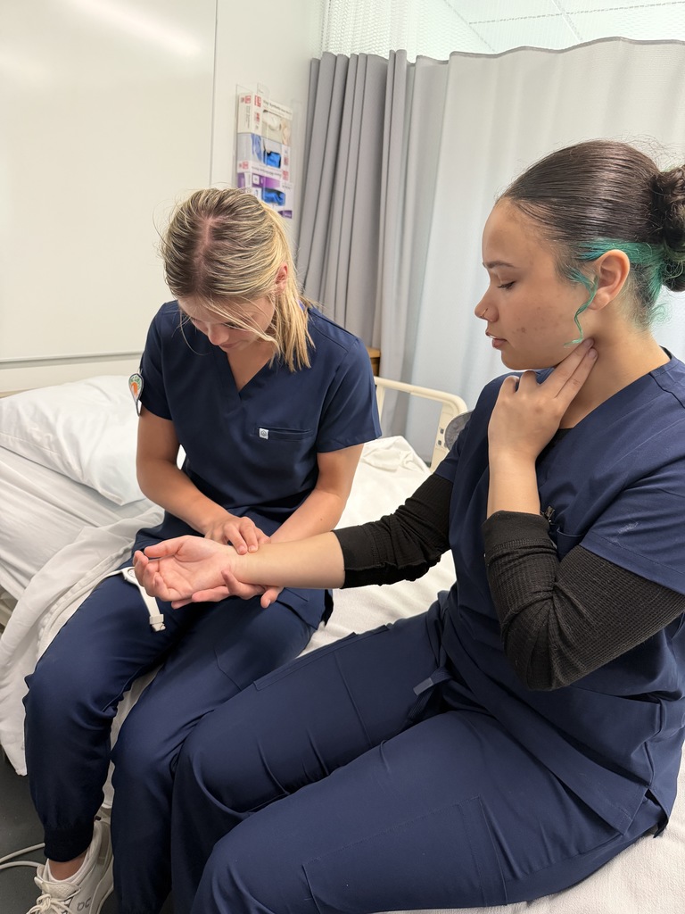 Two women in blue scrubs are seated on a hospital bed. One woman appears to be a patient, and the other is offering care.