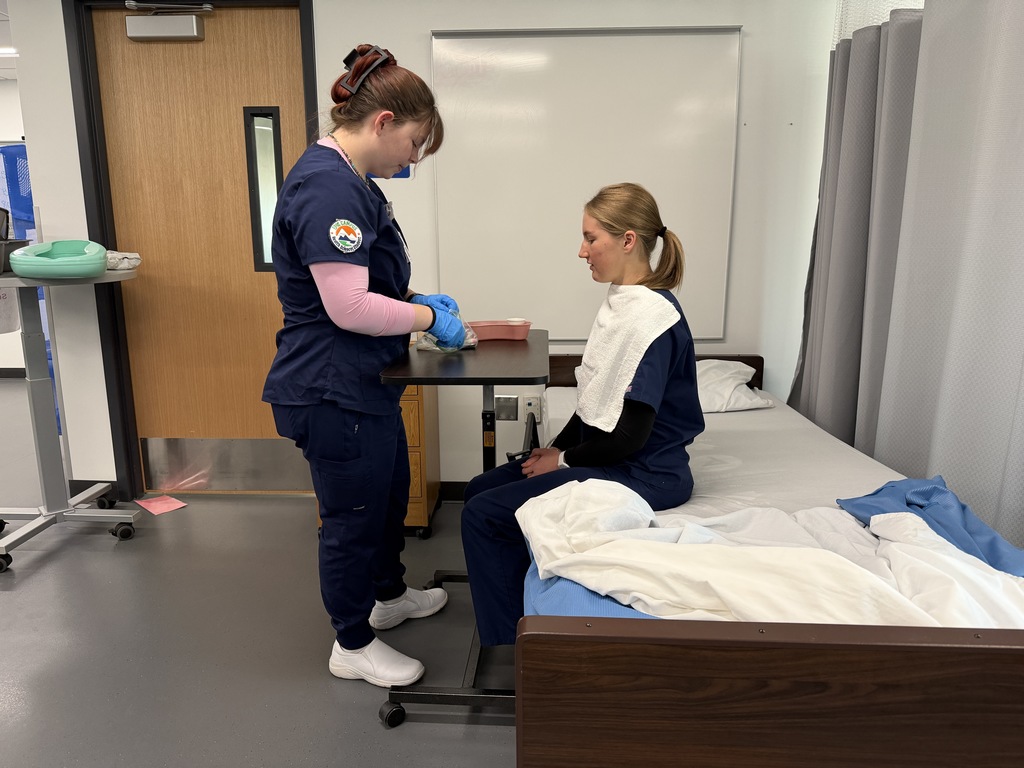 Two women in blue scrubs, one wearing gloves, stand near a bed. The woman in front holds a tray.