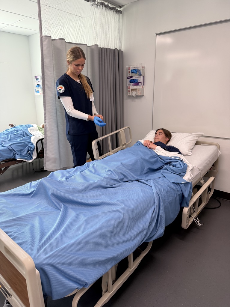 A woman in blue scrubs adjusts a patient in a bed. The patient lies covered with a blue blanket.