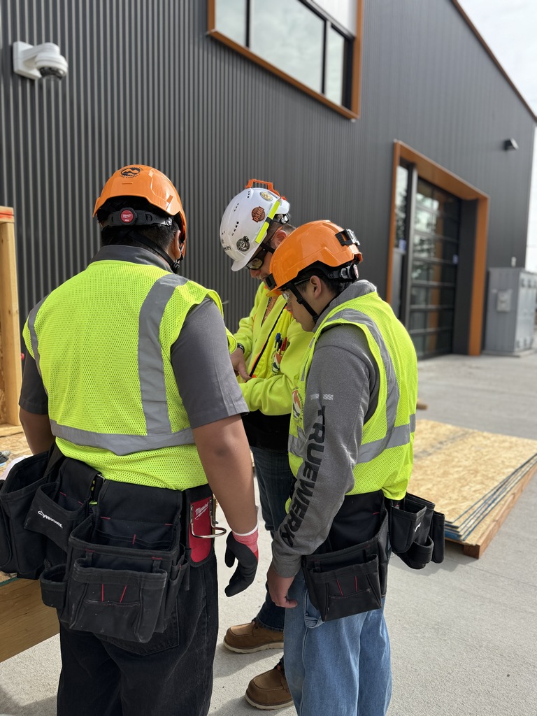 Three people in bright safety gear stand in front of a building. Two wear orange hard hats, and one wears a white hard hat.