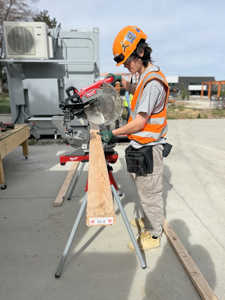 A person in a safety vest and helmet uses a saw to cut a board on a stand.
