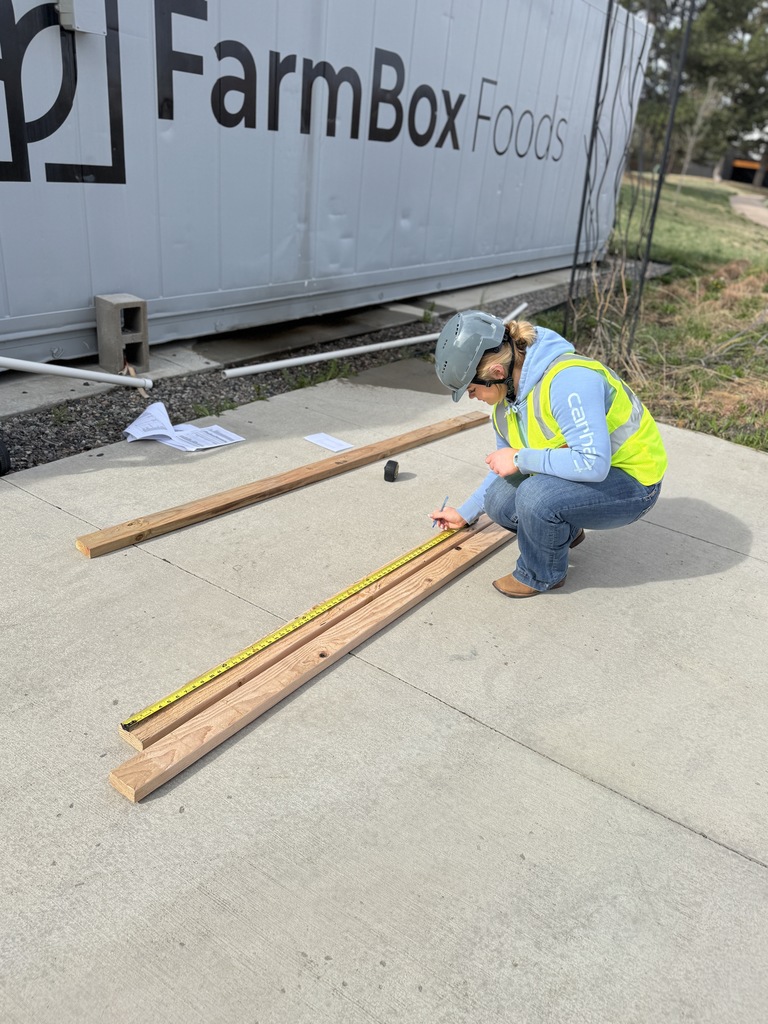 Worker in hard hat and vest measures wooden boards on concrete in front of FarmBox Foods building.