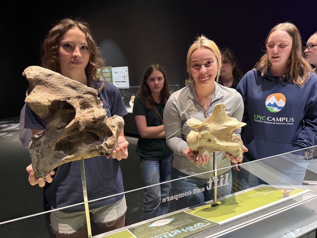 Four women in blue shirts holding fossils in front of a glass display case. A map is on the glass.