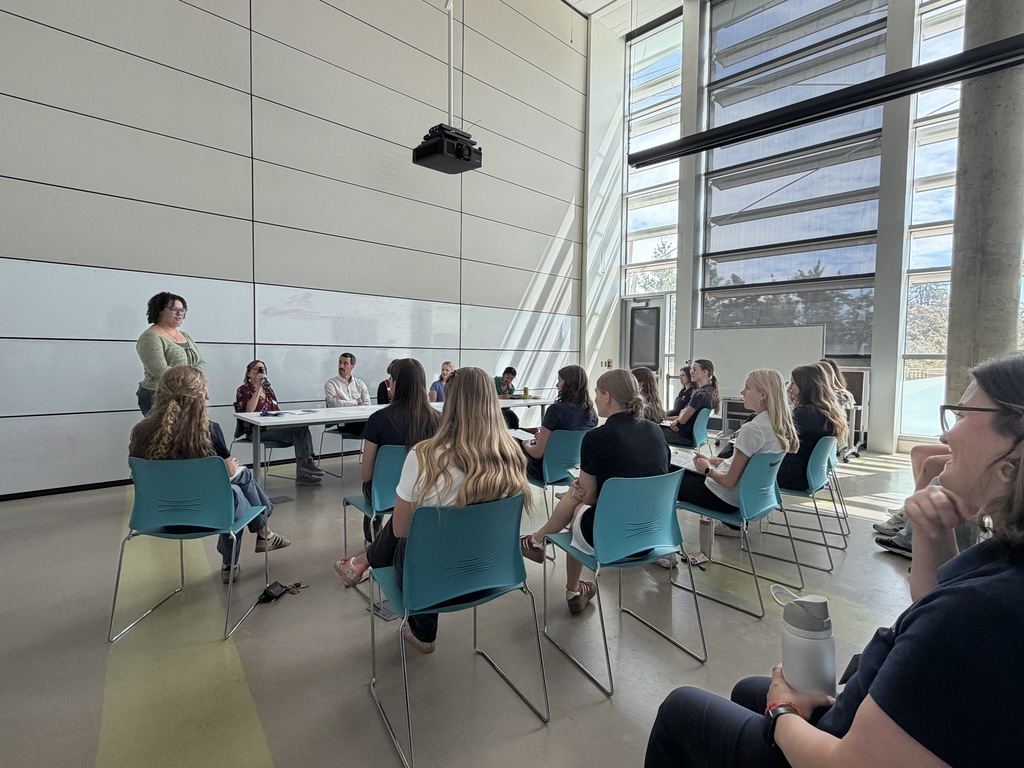 A meeting in a room with large windows. People sit in rows of blue chairs facing the front.
