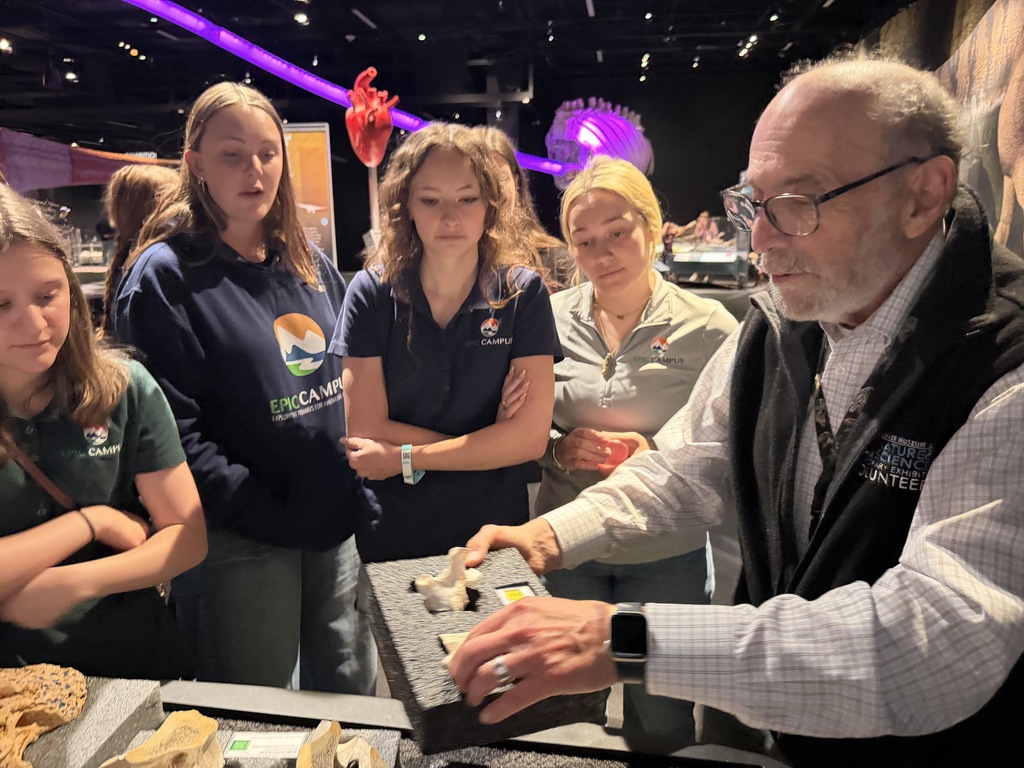 An older man is showing items to a group of young women at an exhibit.