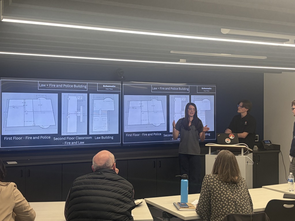 A group of people in a room with large screens. A woman stands before a table speaking.