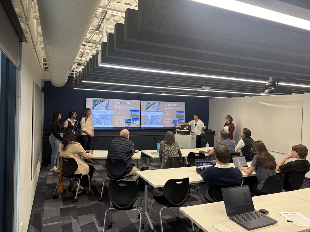 A group of people in a classroom. Some are seated, others standing. A person at a podium faces the audience.