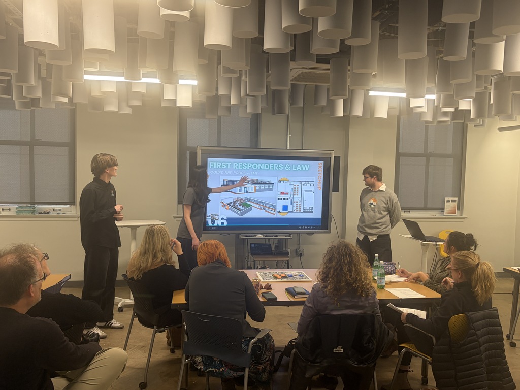 A classroom with several people seated around a table. A large monitor shows a presentation. A person stands at the front of the room.