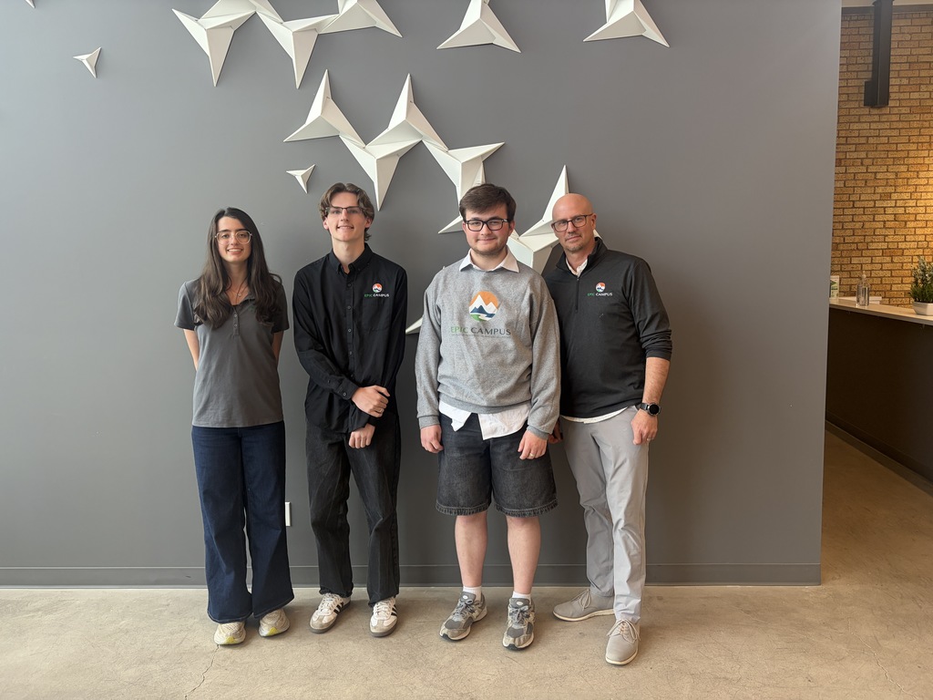Four individuals stand in front of a gray wall with white paper stars. They wear casual clothing and appear to be posing for a photo.