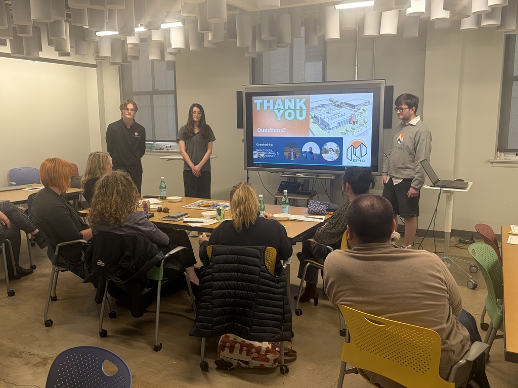 Several people sit around a table, facing a projector screen showing text. Two people stand behind the table.