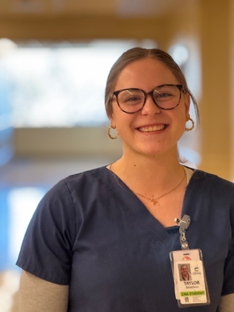 A woman smiles at the camera, wearing glasses, a blue shirt, and an ID badge. She is in a hospital.