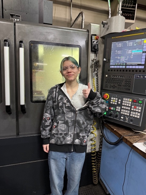 A woman wearing a white top and glasses poses for a photo in a factory next to a large machine.