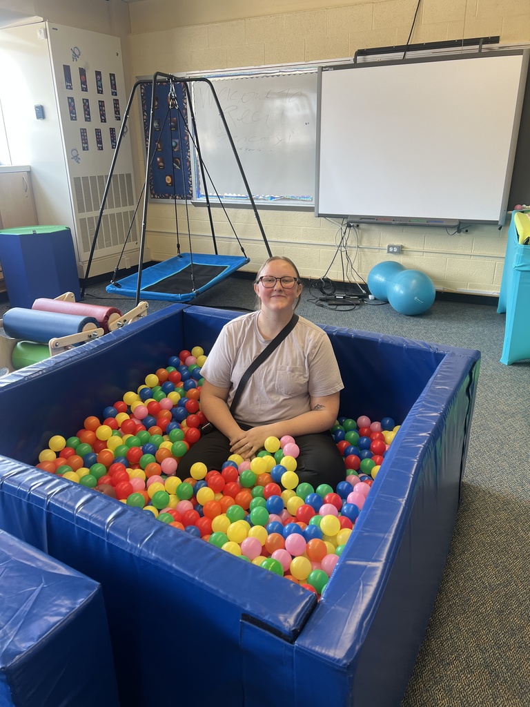A person sits inside a large inflatable ball pit in a room with a whiteboard and various exercise equipment.