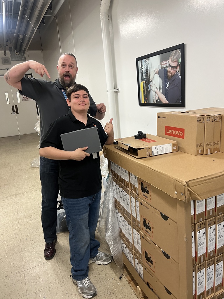 Two people stand near boxes in a room with a framed photo of a man on the wall.