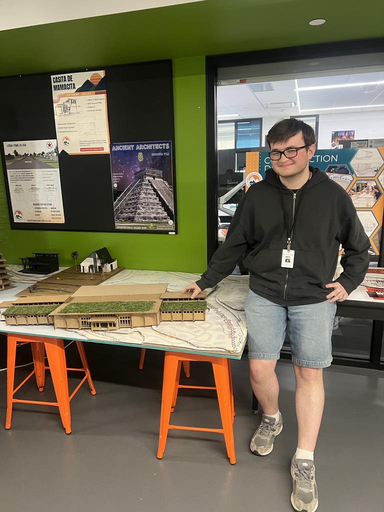 Person stands in front of a desk displaying a small architectural model. A green wall with posters in the background.