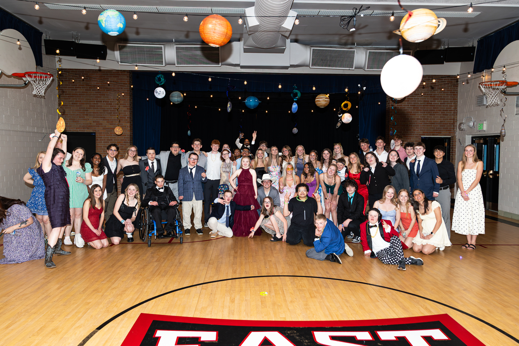  A large group of approximately 50 young adults poses for a group photo on a wooden basketball court during a formal or semi-formal event, likely a prom or dance. The setting is a gymnasium decorated with a space theme, featuring hanging paper lanterns shaped like planets (including Saturn and Earth) and string lights.