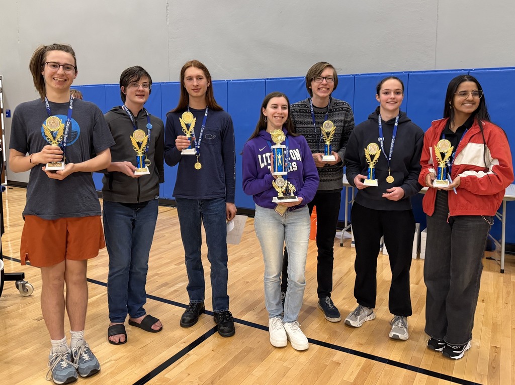A group of students in a gym, holding trophies. A blue wall and wooden floor are in the background.