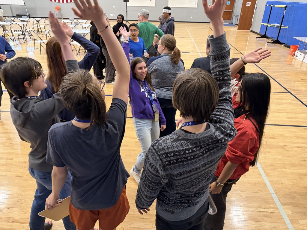 Several people stand in a gymnasium, raising their hands in a circle. Some wear casual clothing; others wear name tags.
