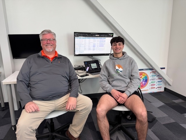 Two people in a room, one in a gray hoodie and the other in glasses, seated in front of a desk.