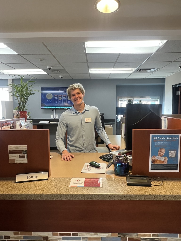A person stands behind a counter in a room with a desk, computer monitor, and plants.