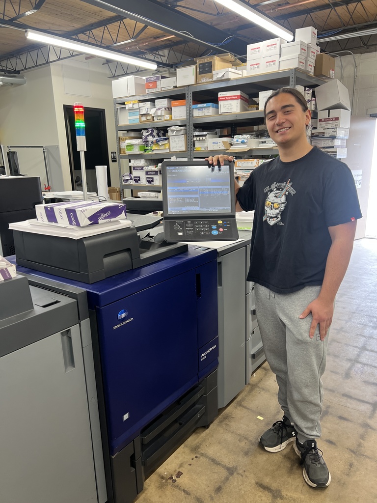 A person wearing a black t-shirt and grey pants stands next to a blue printer. Behind them are shelves with boxes.
