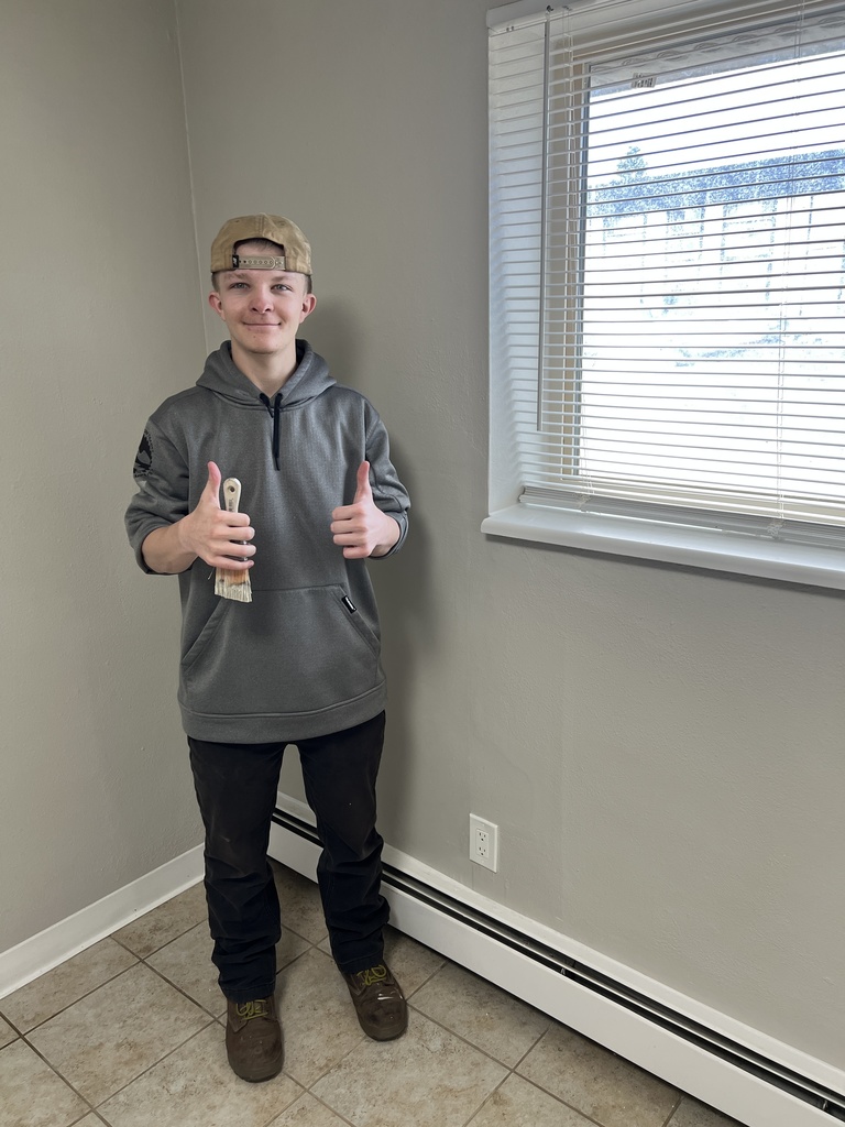 Man in a gray hoodie giving a thumbs up in a room with light-colored tiles and a window.