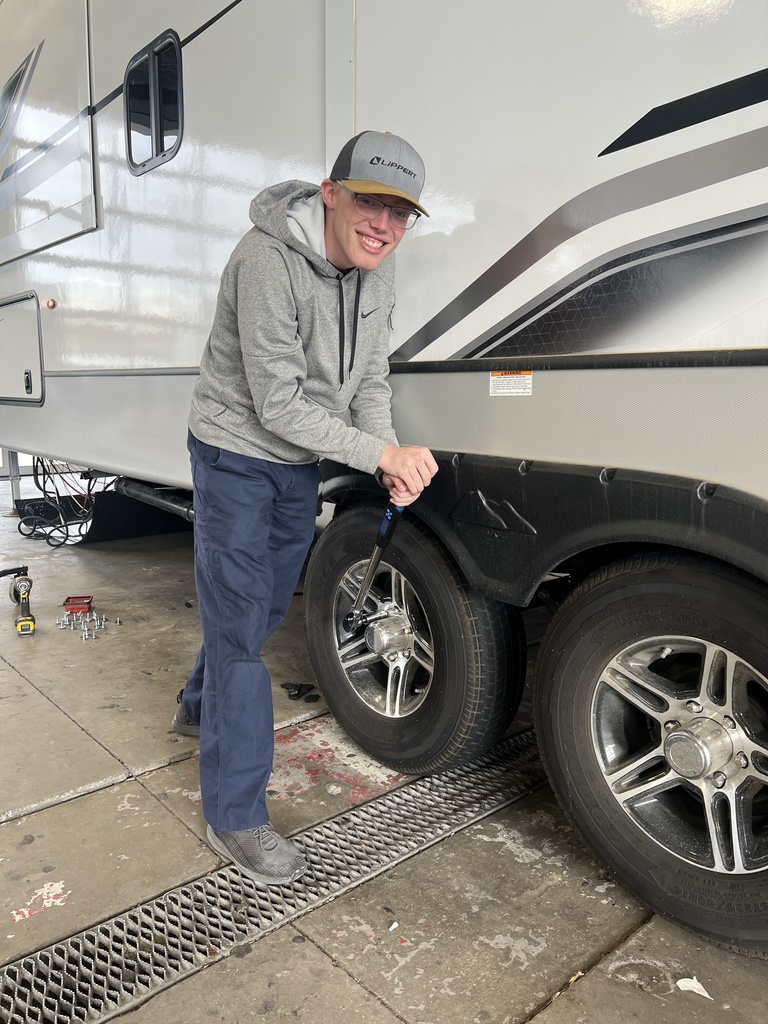 A man with a hat and glasses holds a tire iron by the tire of a recreational vehicle.