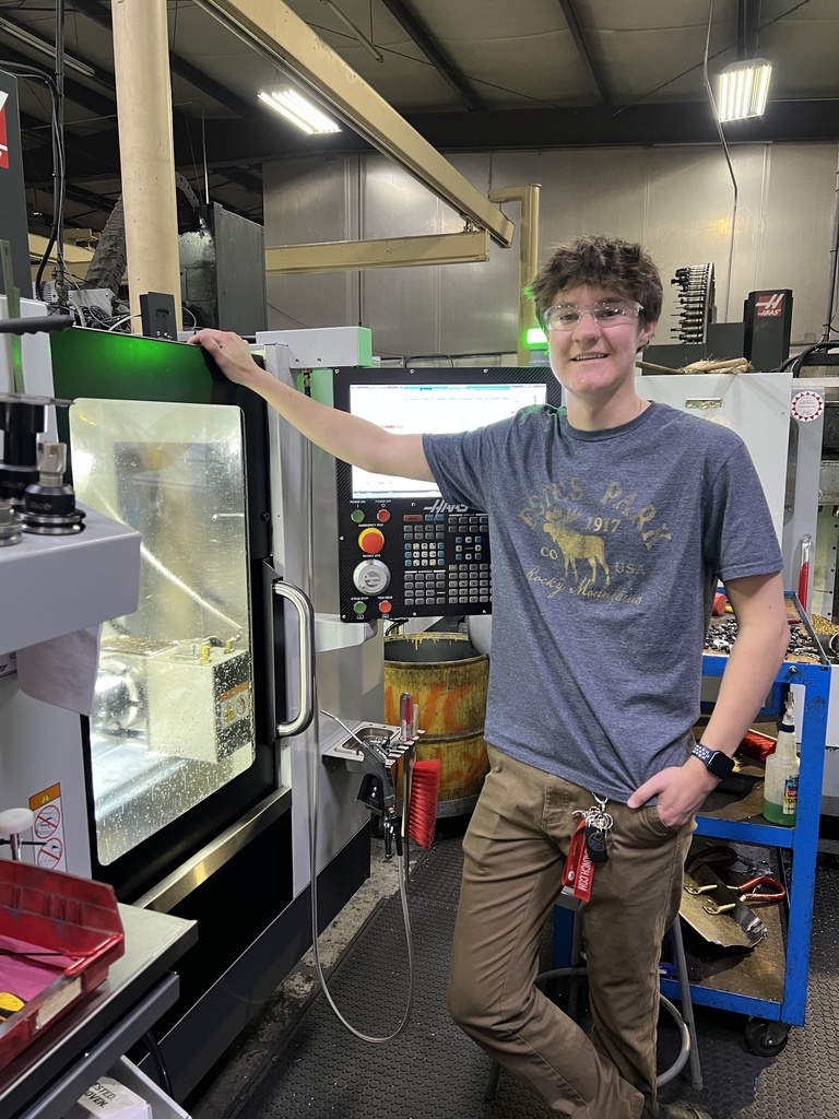 A man in a gray t-shirt and brown pants stands by a large green machine in an indoor factory.