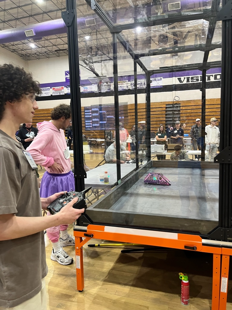 A person holds a remote control and operates a robot inside a large, transparent cage.