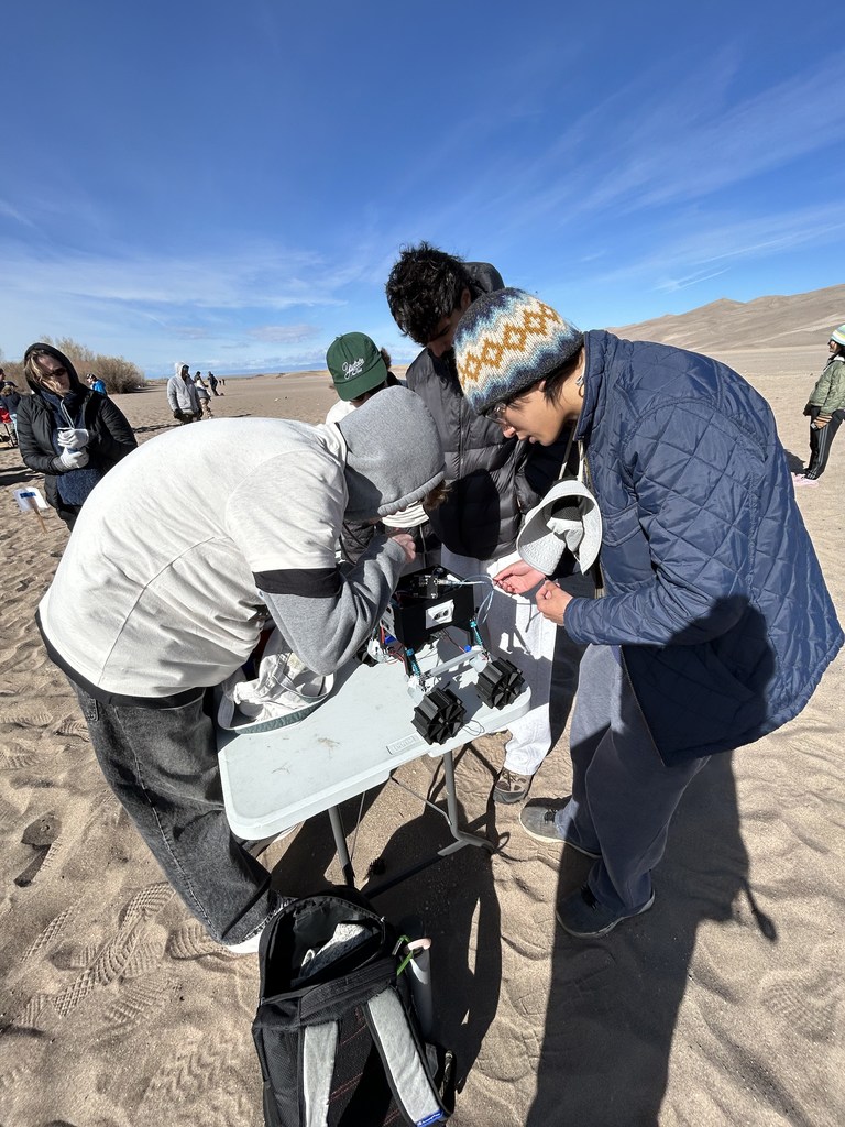 People in winter wear examine objects on a table under a clear sky in a desert.