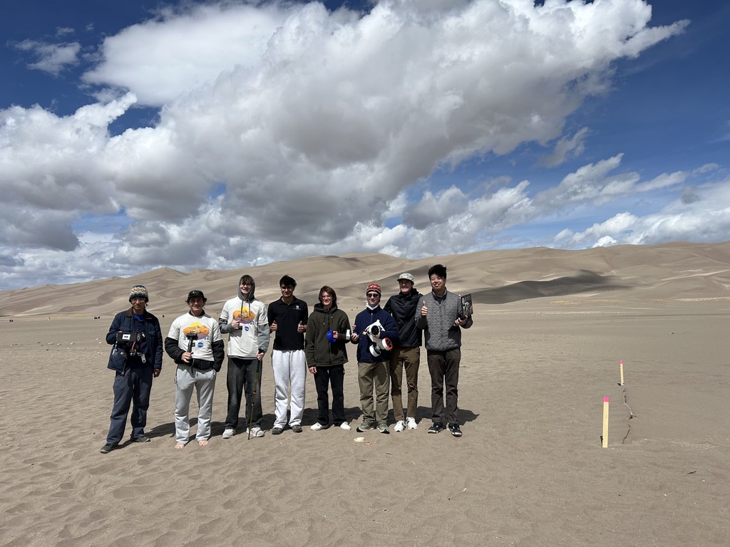 Nine people stand in a sandy desert with dunes in the background, under a partly cloudy blue sky.