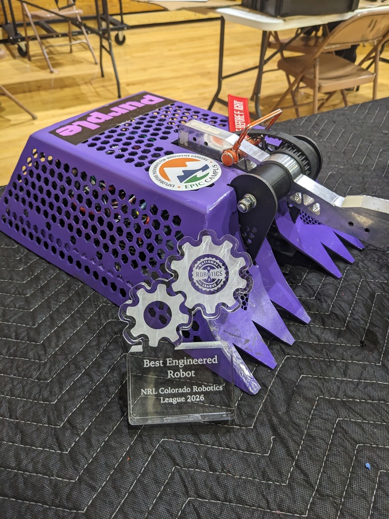 A purple robot on display, featuring a plaque that reads "Best Engineered Robot" and a logo. A table with a black cover is in the background.