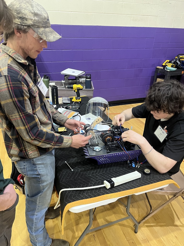 Two people working together at a table with tools and equipment on it. A purple wall behind them.