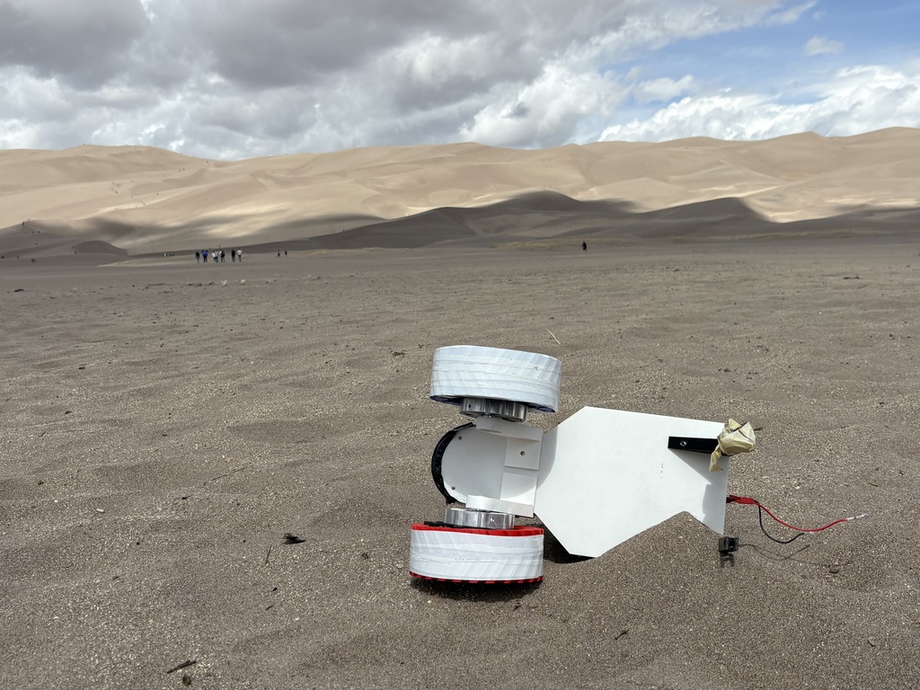 A small, white, wheeled robot with round wheels rests in a sandy, desert landscape with distant dunes under a partly cloudy sky.
