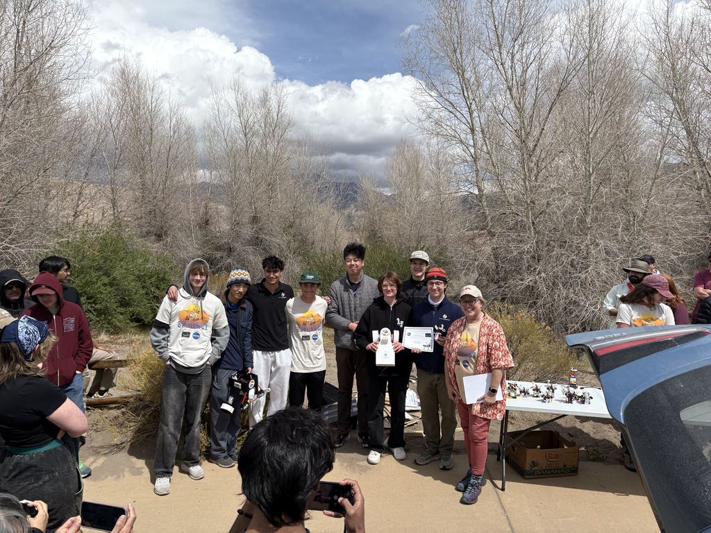 People gathered in a line, some holding signs, standing on a dirt path with trees and mountains in the background.