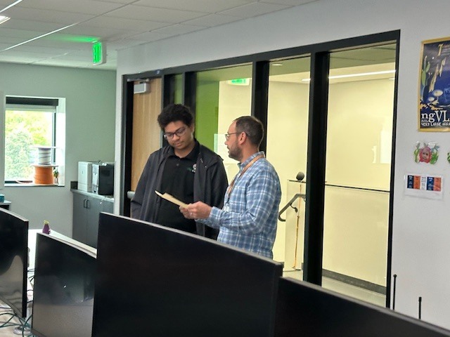 Two men in a room with glass walls and monitors. One man reads from a paper while the other listens.