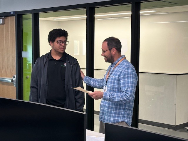 Two men are indoors, one holding paper, the other wearing glasses. A glass wall is behind them, with a door to the left.