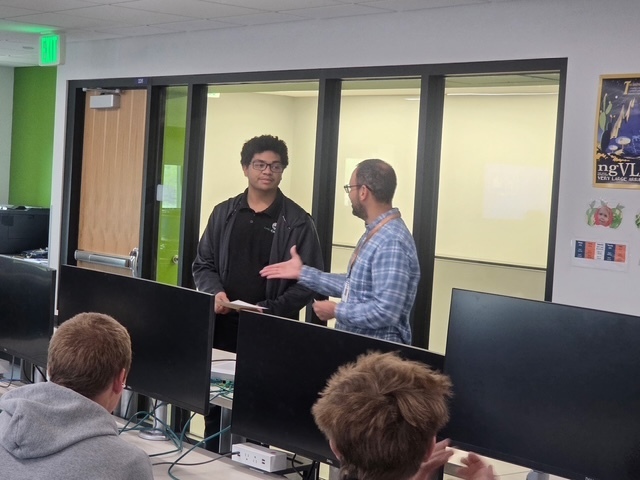 A group of people sit at computers in a classroom, two men in front talk with each other.