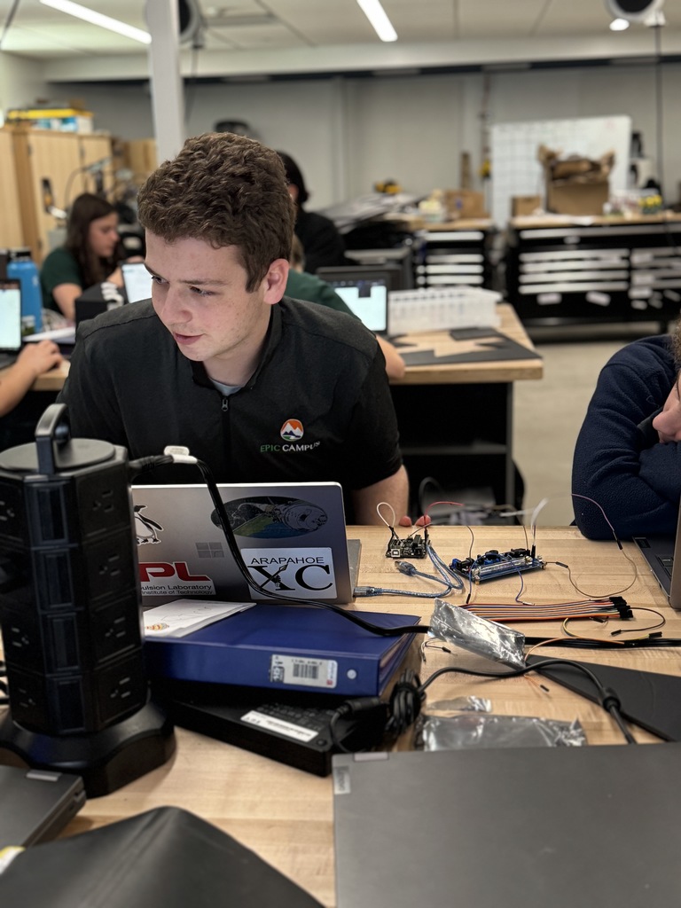 A man in a black shirt works on a laptop at a desk with various devices.