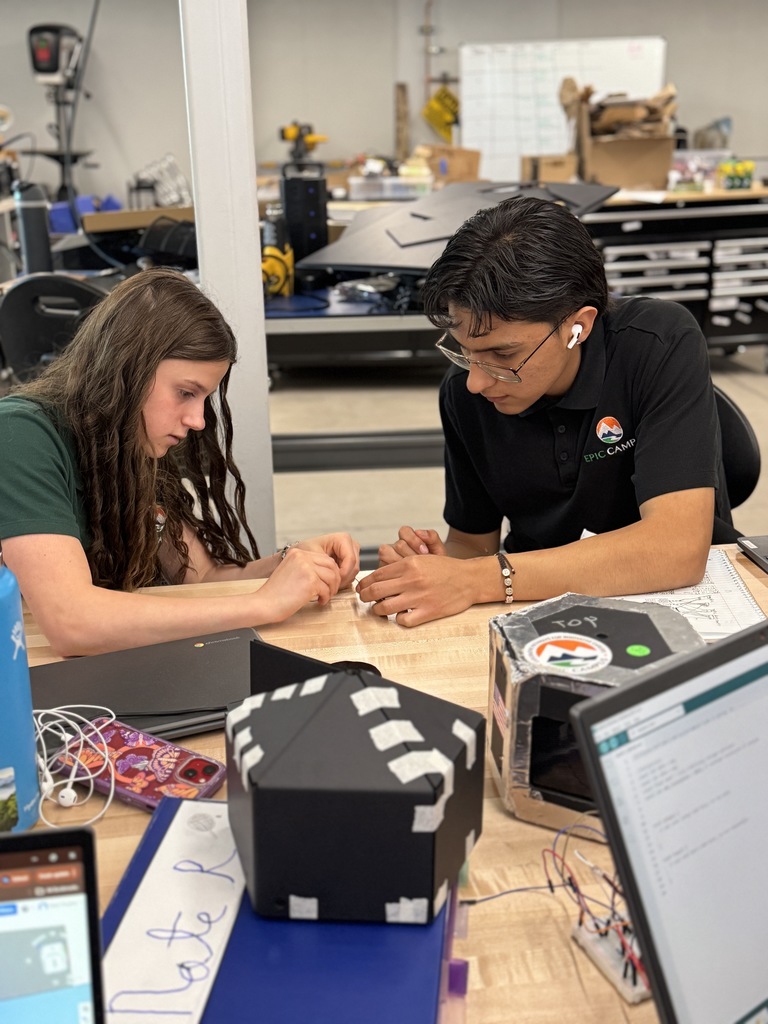 Two people work together on a project in a workshop, one sitting at a desk with a laptop.