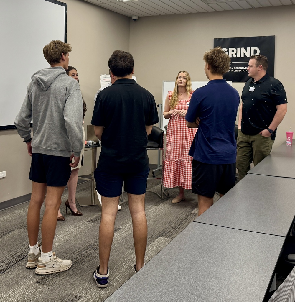 People standing in a meeting room. Two men in front, two women in back. One man wears a watch, woman in pink dress.