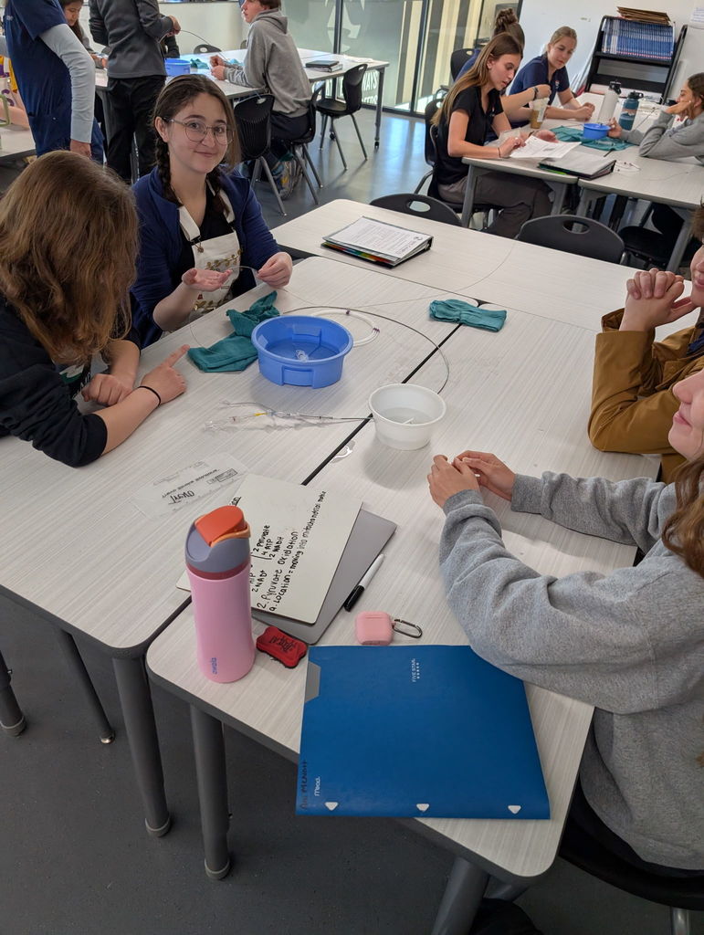 Several students sit around a table, working on an art project. One holds a piece of paper and another holds a water container.
