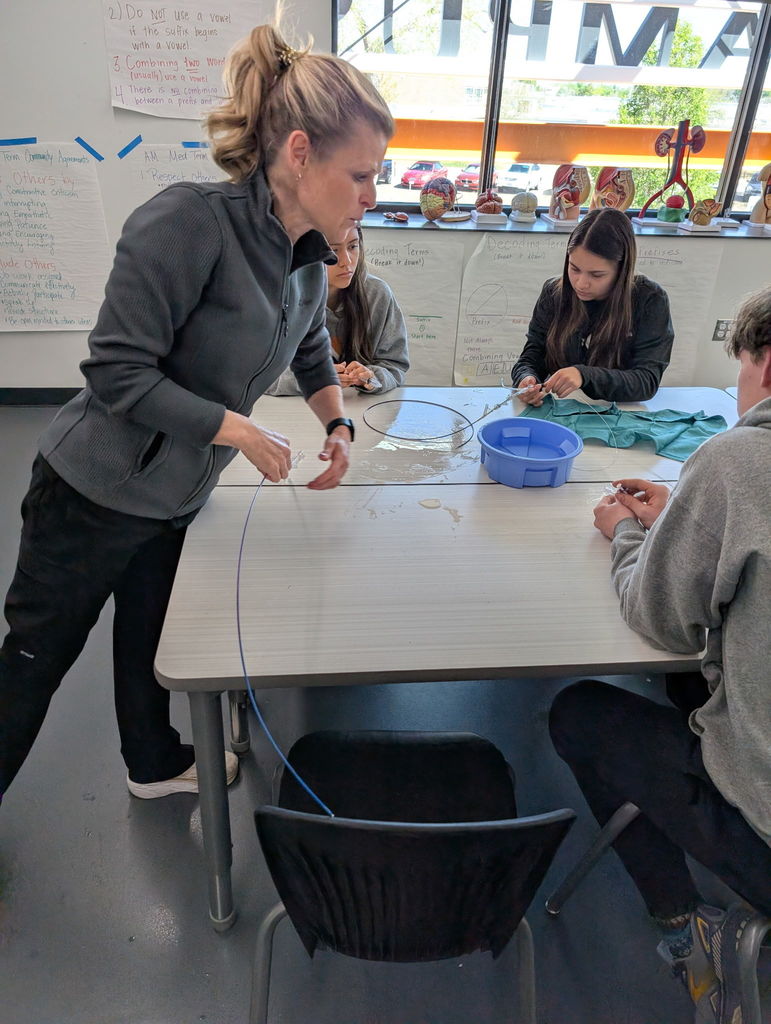 Three individuals sit around a table, engaged in a hands-on activity with a wire. One person stands, holding the wire, while others work on it.