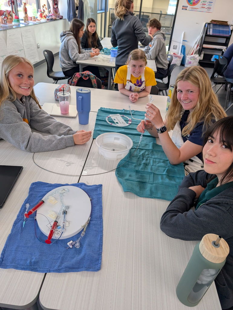 Four people sit at a table, with two women smiling, one wearing a yellow apron. Art materials and a blue cloth are on the table.
