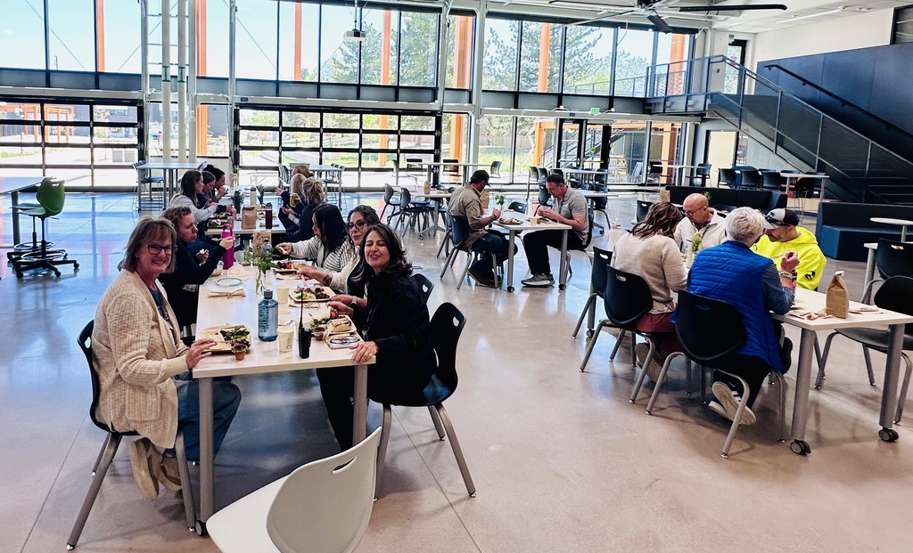 A group of people sitting at tables in a large, open space with glass windows and a staircase.