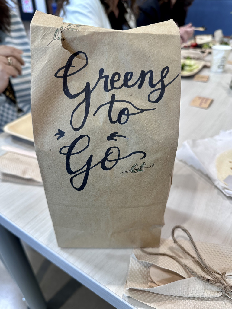 A paper bag with "Greens to Go" written in blue on a table with a cloth napkin, a plate, and a cup.