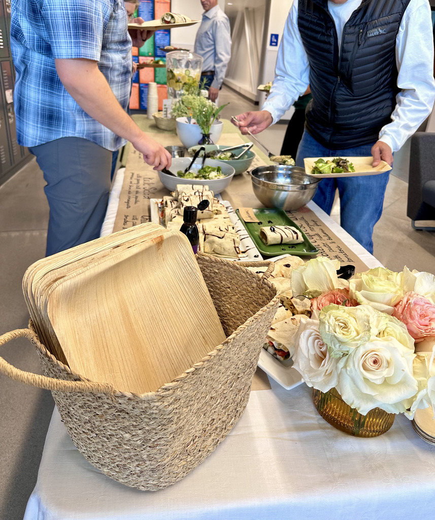 A table with a white tablecloth holds a basket, flowers, plates, and a bowl. People prepare to serve food.