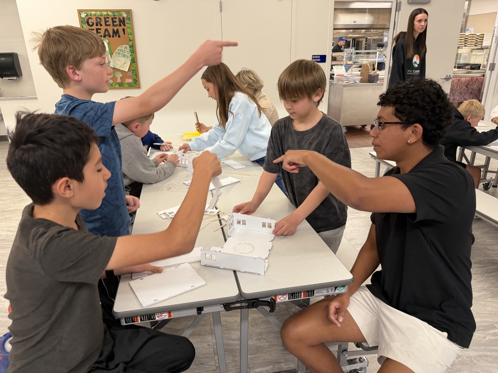 Several children are sitting at a table inside a classroom, engaged in an activity. A woman is standing in the background.